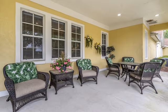 Outdoor patio seating with wicker chairs, tables, and potted plants beside a yellow wall.