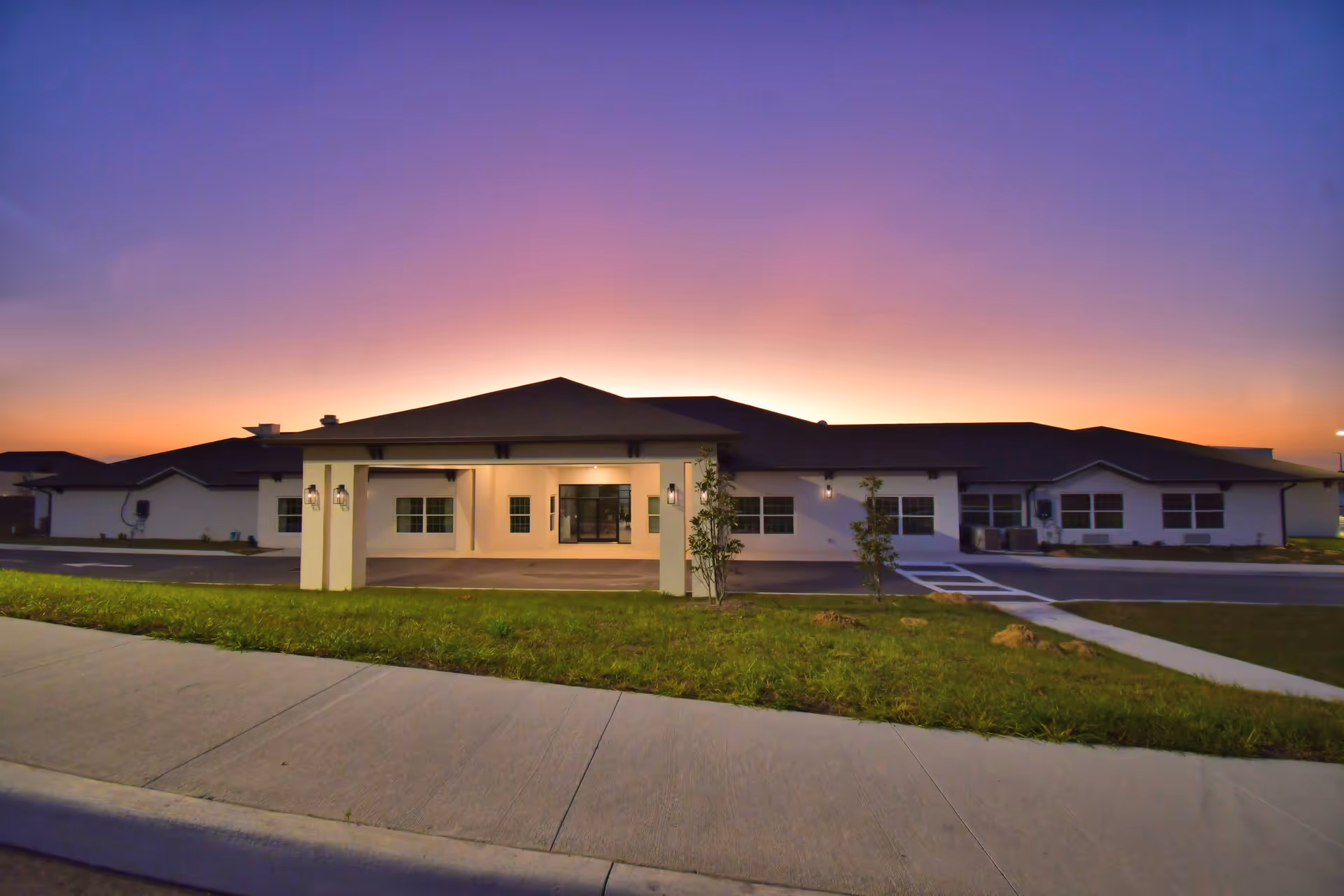 Single-story senior living facility with a covered entrance, parking, and sunset sky.