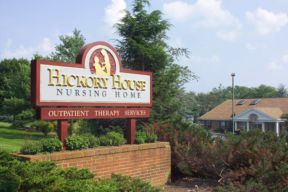 A landscaped entrance sign reading "Hickory House Nursing Home" with shrubs, trees and the facility building in the background.