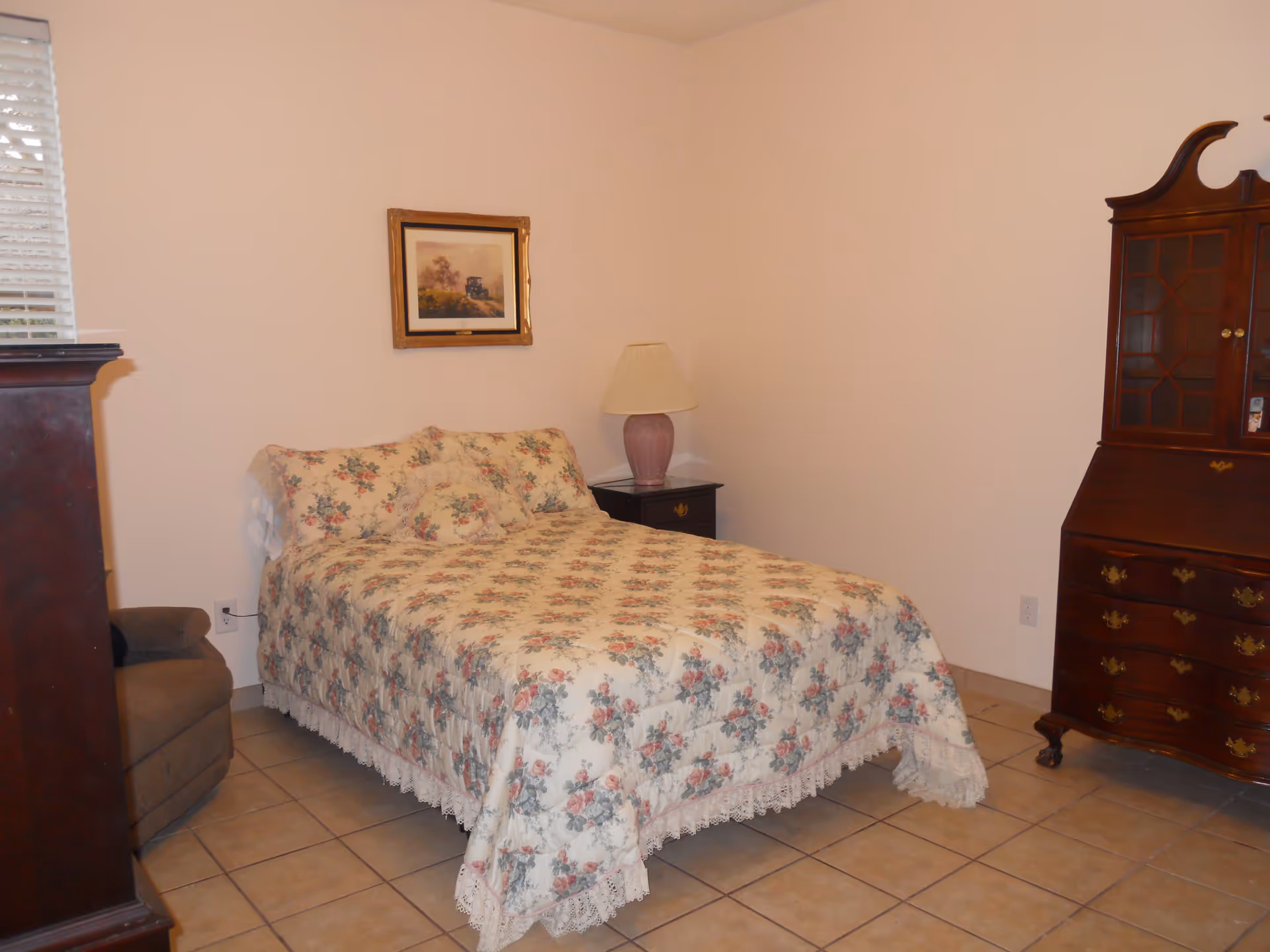 A bedroom with a bed covered in a floral patterned bedspread, a nightstand with a lamp, a framed picture on the wall, a wooden dresser with glass doors, and a cushioned chair in the corner. The floor is tiled and the walls are plain white.