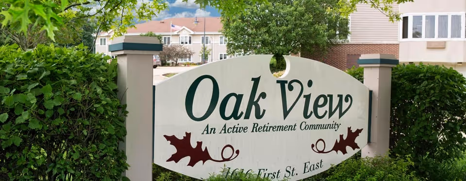 A wooden entrance sign that reads "Oak View An Active Retirement Community" set among bushes with the facility building in the background.