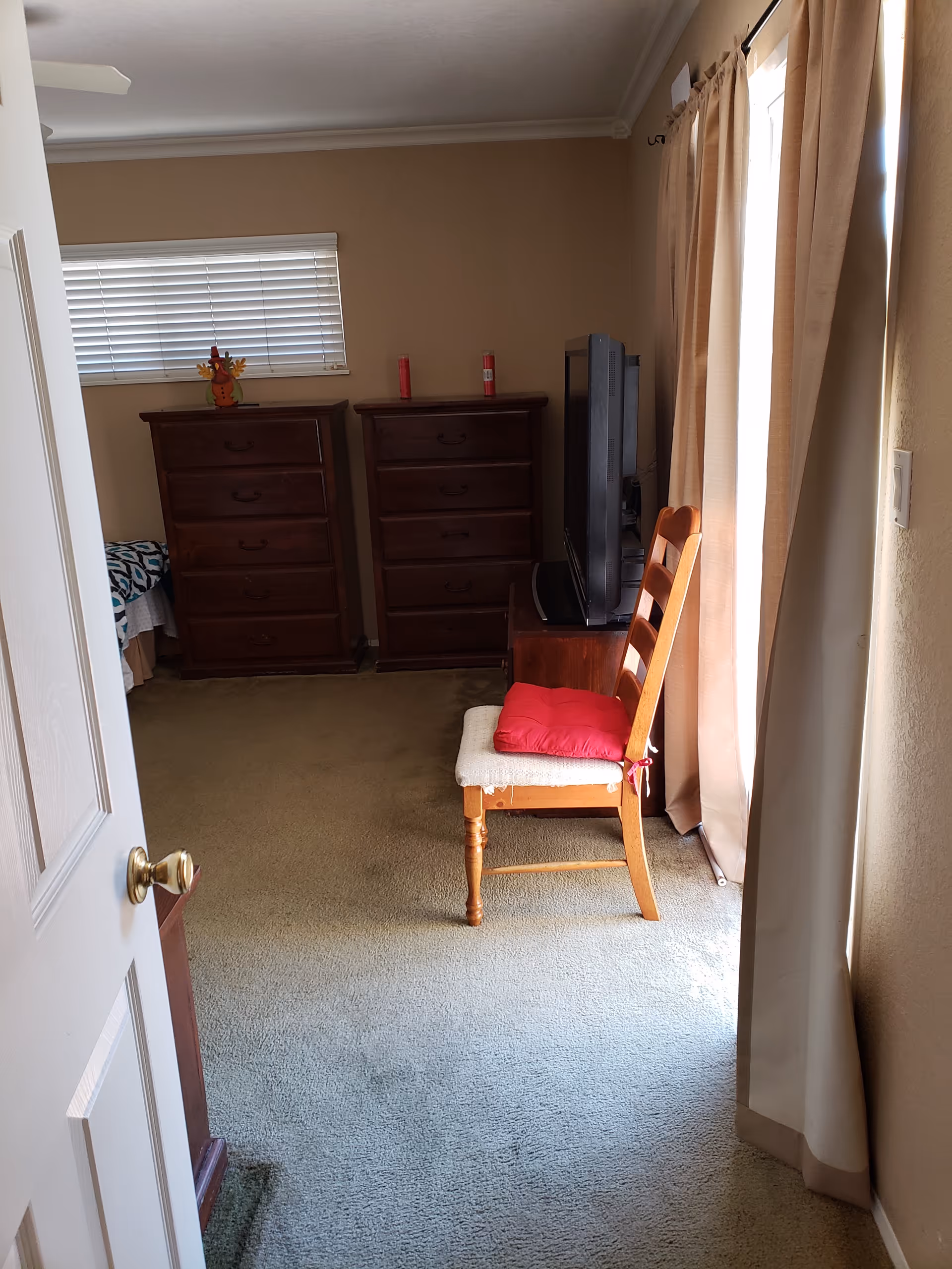 Small bedroom with two dressers, a chair, TV, and curtains in a senior living facility.