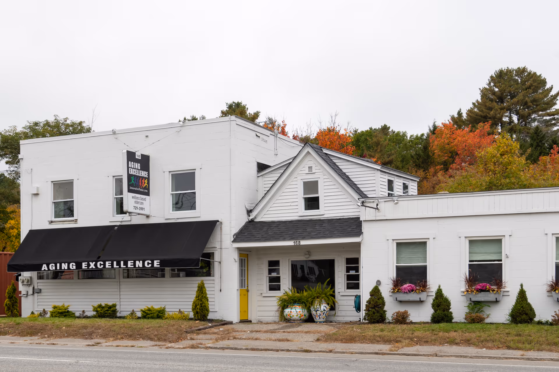 White senior living facility building with a black awning labeled 'Aging Excellence' and a front entrance.