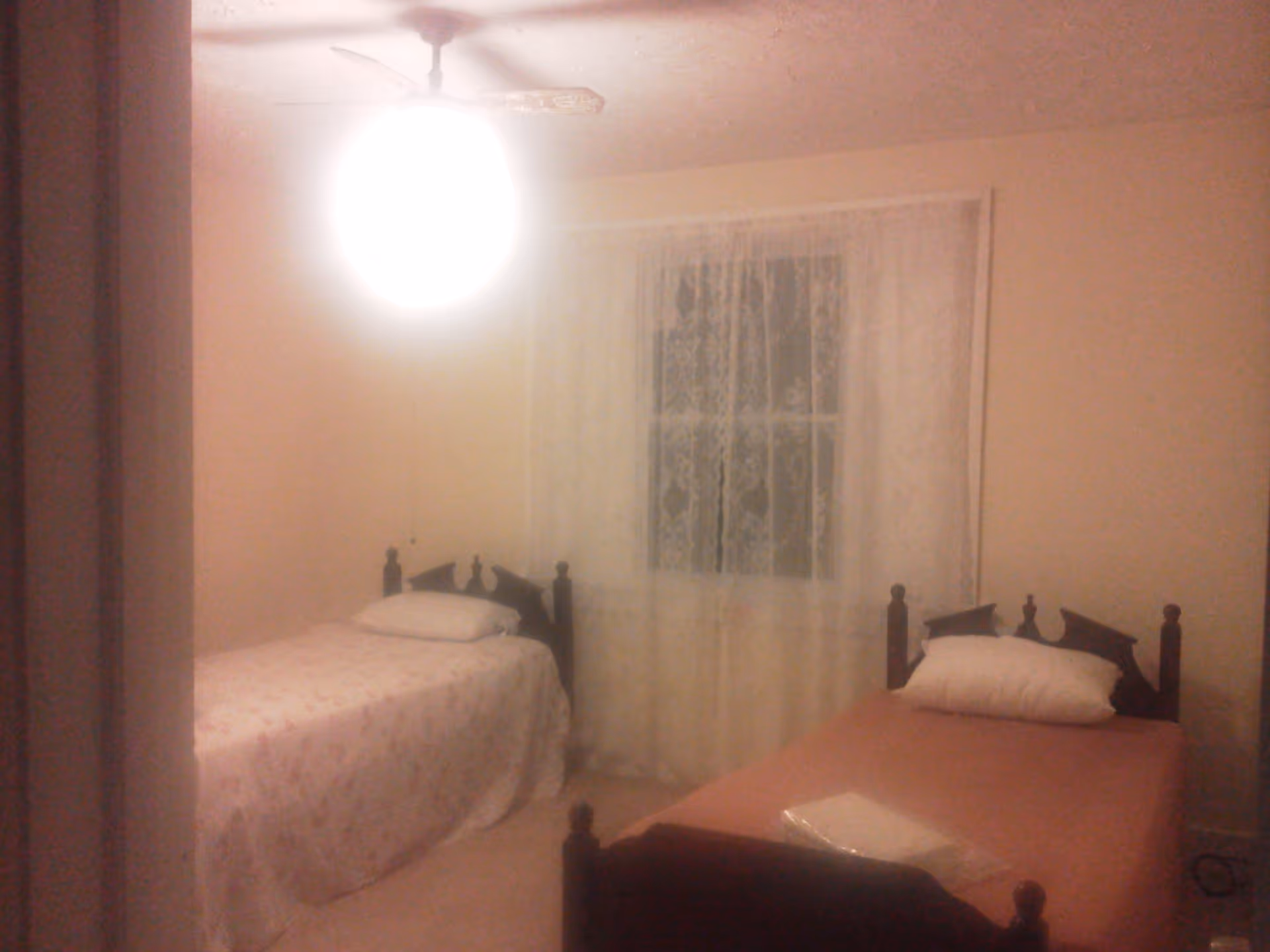A small bedroom with two single beds placed parallel to each other. Each bed has a wooden headboard and a pillow. One bed has a pink bedspread and the other has a floral-patterned bedspread. A window with lace curtains is centered on the wall between the beds. A ceiling fan with a bright light is on the ceiling.