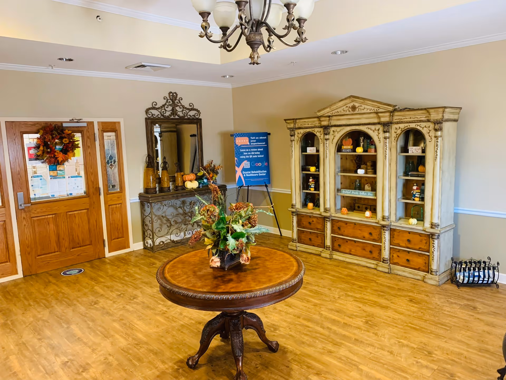 A bright reception lobby with a round wooden table topped with a floral arrangement, decorative cabinet shelving, and double wooden entry doors with a wreath.