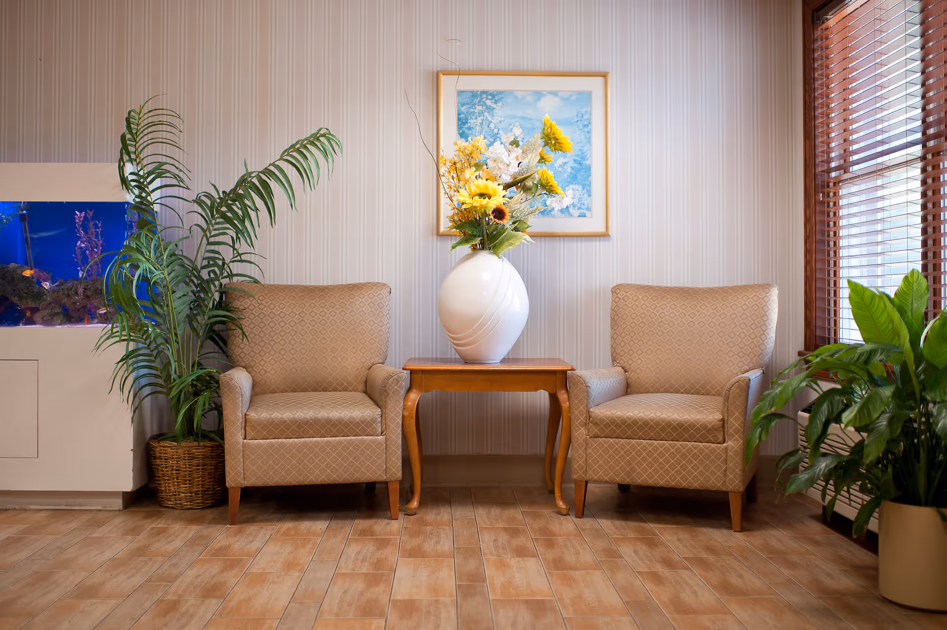 A cozy seating area with two beige patterned armchairs separated by a wooden side table holding a white vase filled with yellow and white flowers. There is a framed painting of white flowers on a blue background hanging on the wall behind the table. To the left, there is a tall green potted plant next to a built-in aquarium with colorful fish. On the right, a window with wooden blinds lets in natural light, and another green potted plant is placed near the window. The floor is covered with light brown tiles.