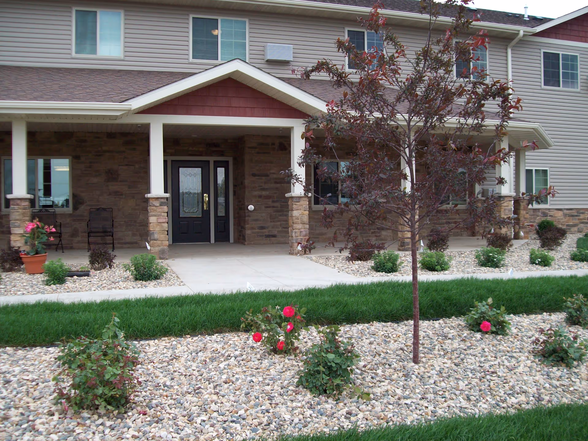 Front entrance of a two-story senior living building with a covered porch, stone columns, and landscaped rock beds with small trees and roses.