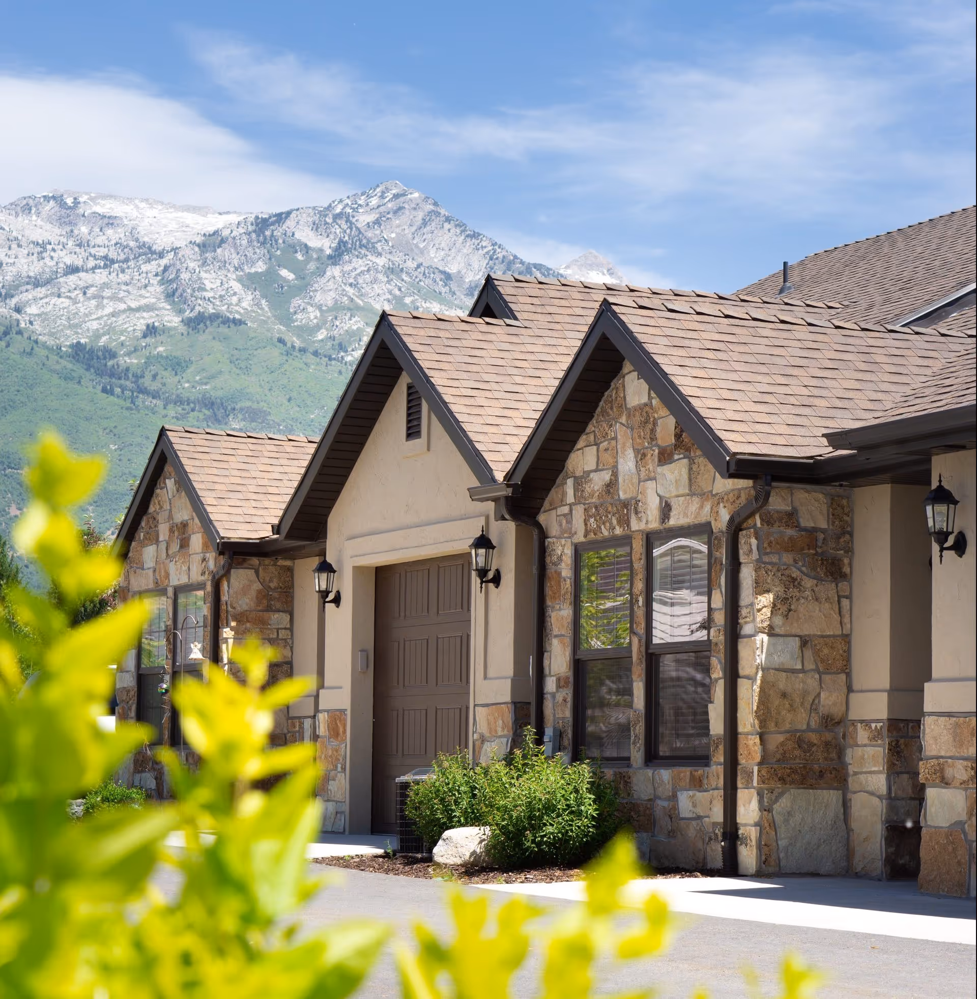 Front exterior of a stone-faced single-story building with gabled roofs, a garage door, lanterns, shrubs, and mountains in the background.