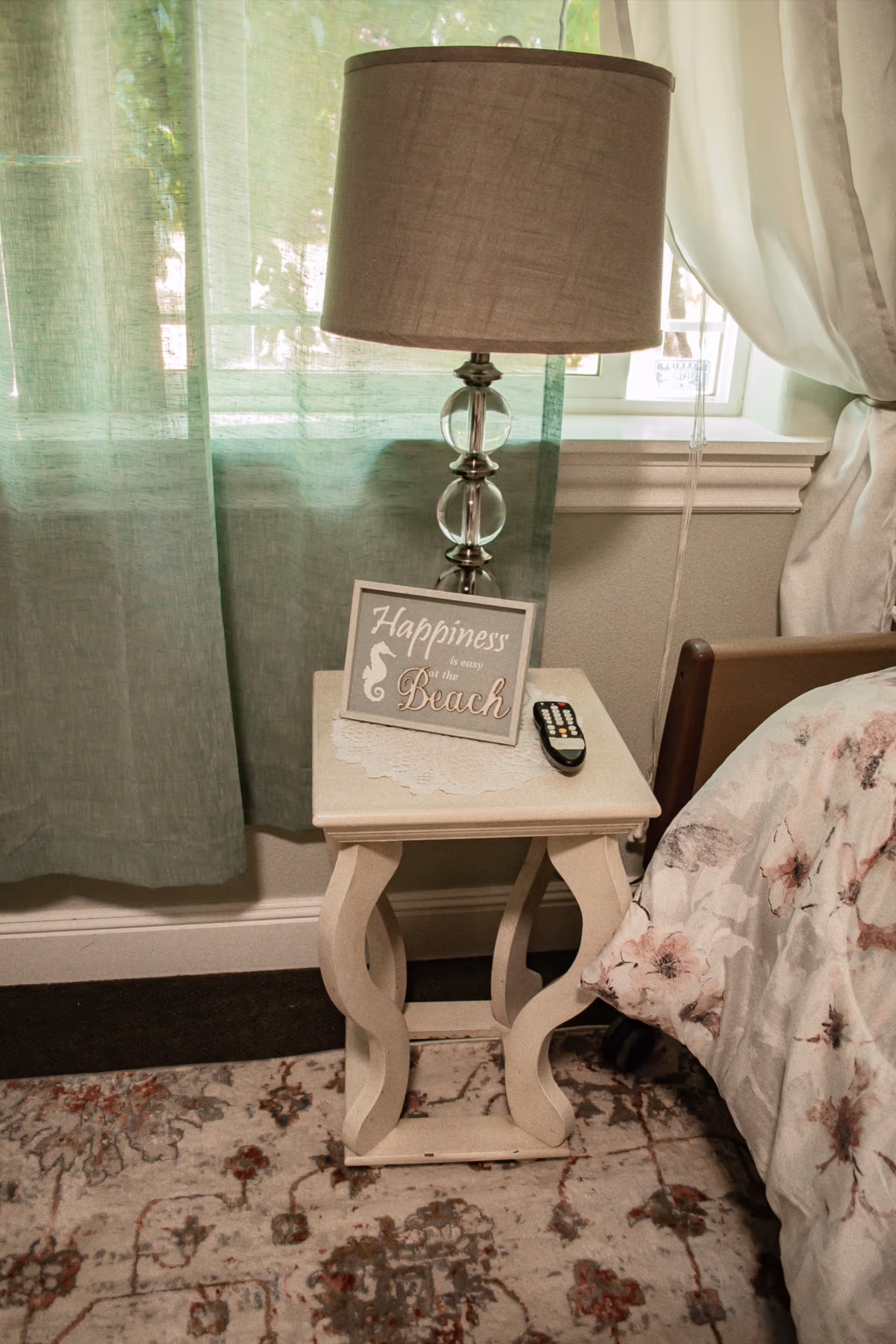 A bedside table with lamp, remote, and decorative sign beside a floral bed.
