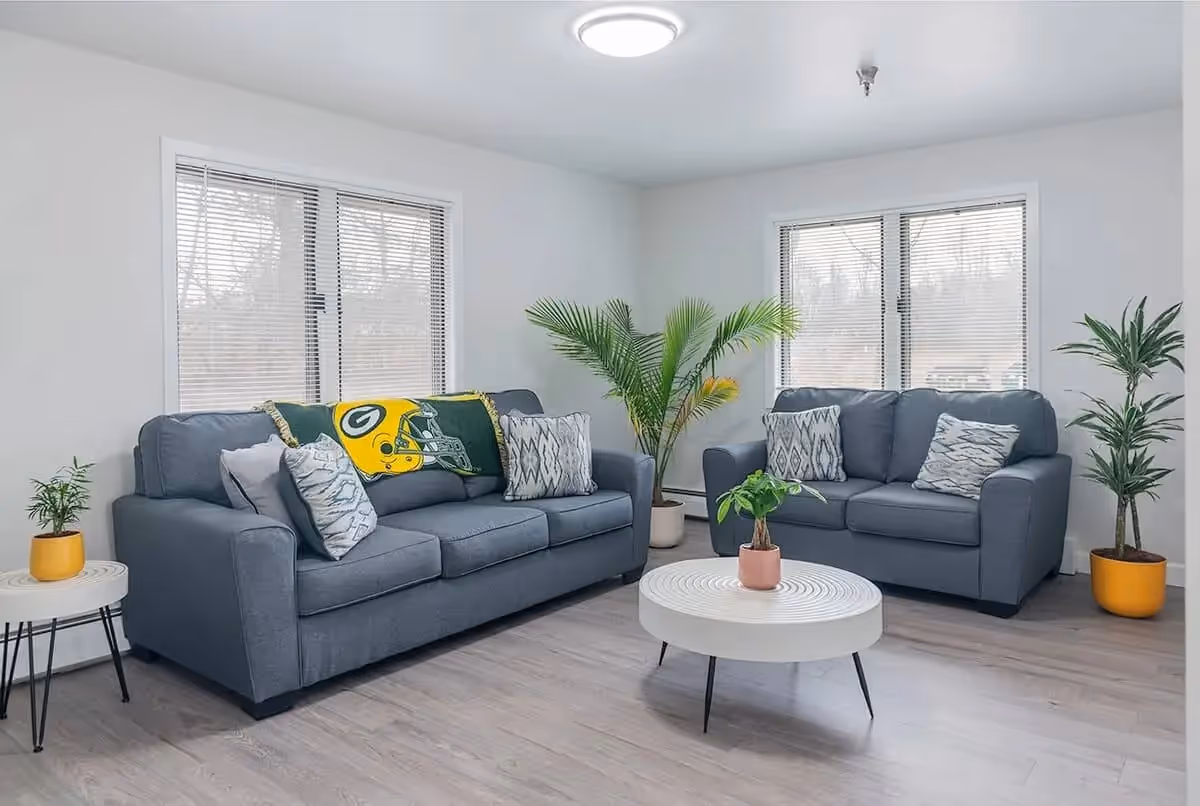 Bright sitting area with two gray sofas, patterned pillows, plants, and a round white coffee table in a modern living room.