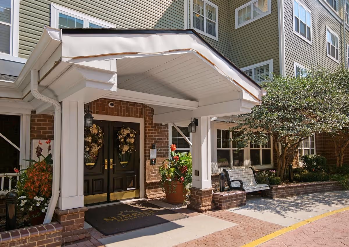 Covered main entrance to a senior living building with double doors, wreaths, potted plants, and a bench.