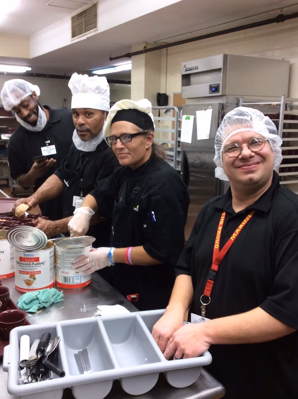 Four kitchen staff members wearing hairnets and chef hats are preparing food in a commercial kitchen. They are standing behind a stainless steel counter with large cans of butterscotch pudding and various kitchen utensils. One person is smiling at the camera while the others are focused on their tasks.