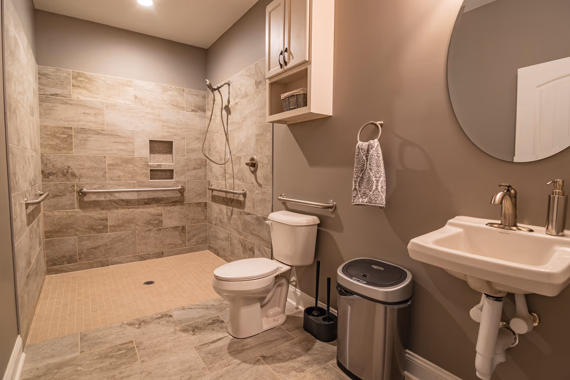 Accessible bathroom with tiled shower, grab bars, toilet, sink, and round mirror.