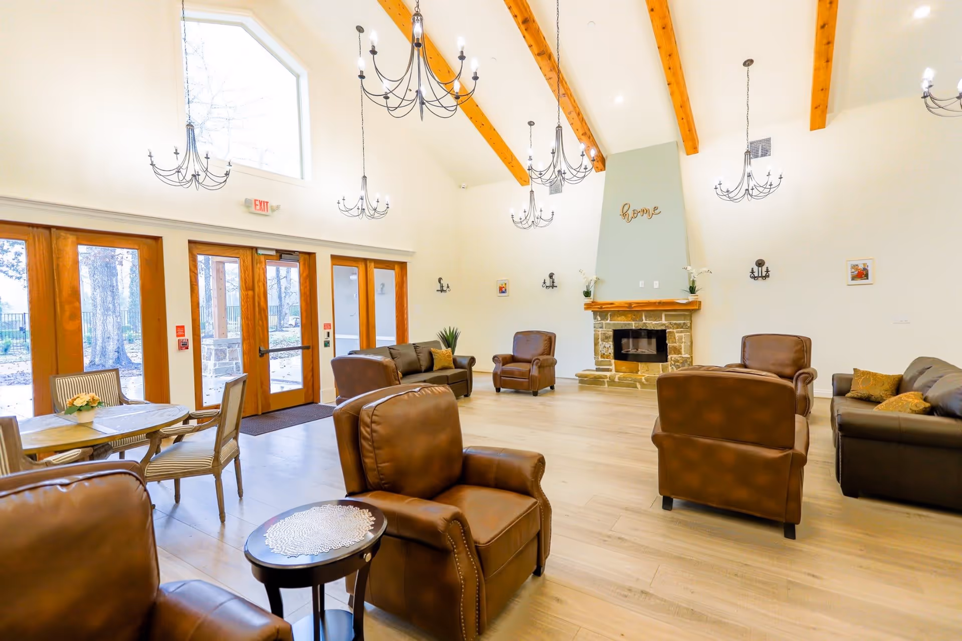 Spacious memory-care community lounge with vaulted beamed ceiling, chandeliers, a stone fireplace, leather armchairs and sofas arranged around tables near large wooden doors and windows.