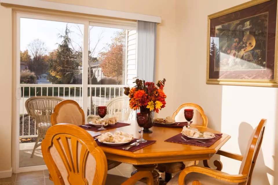 A dining area with a wooden table set for four, featuring burgundy placemats, beige napkins, and red wine glasses. A floral centerpiece with autumn-colored flowers is in the middle of the table. Behind the table is a sliding glass door leading to an outdoor patio with wicker chairs. A framed painting hangs on the wall to the right.