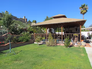 A sunny outdoor area featuring a wooden gazebo with a shingled roof surrounded by green grass, plants, and trees, including a tall palm tree in the background under a clear blue sky.