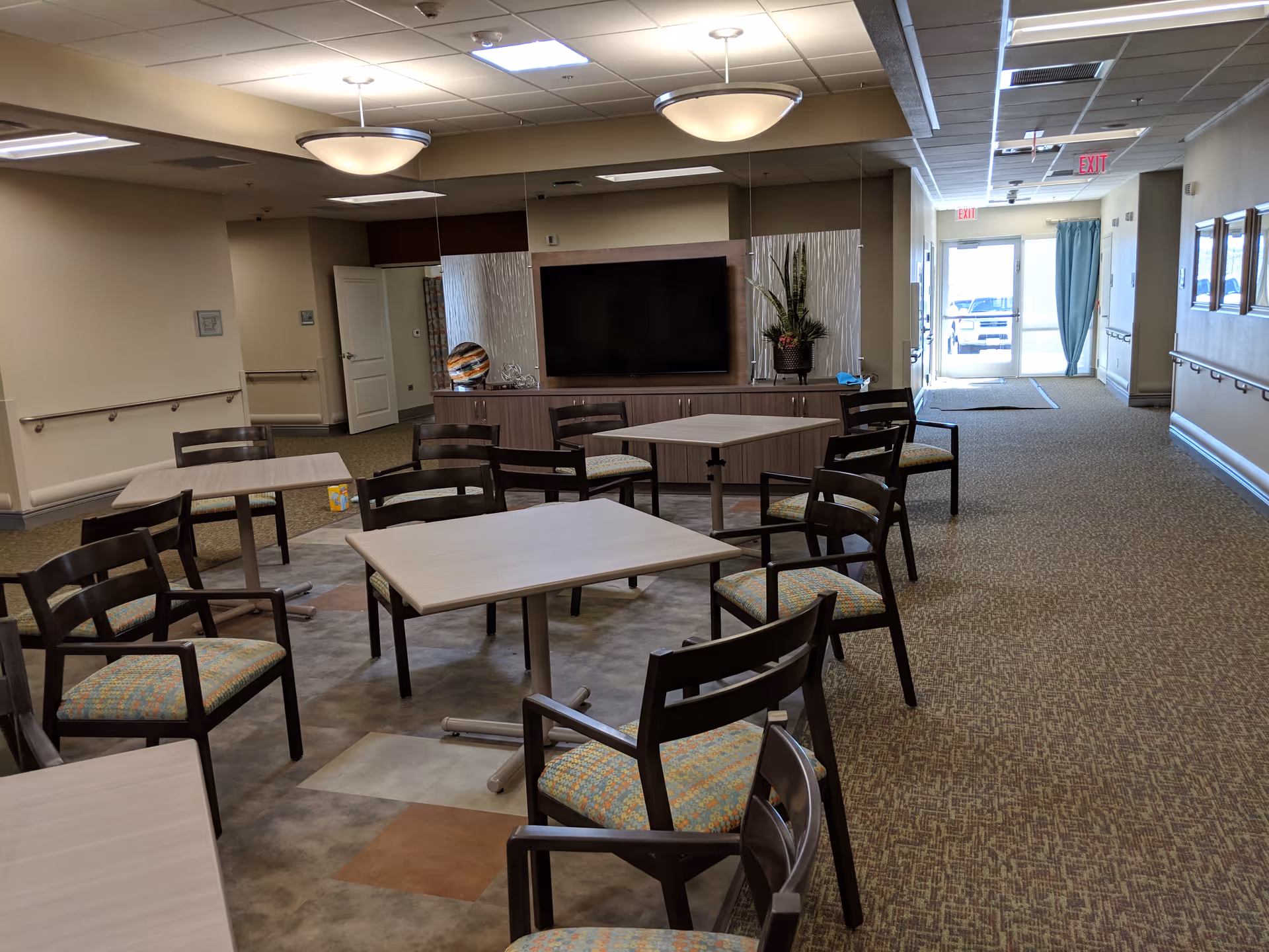 Senior living facility common area with multiple tables and chairs, TV, and hallway exit.