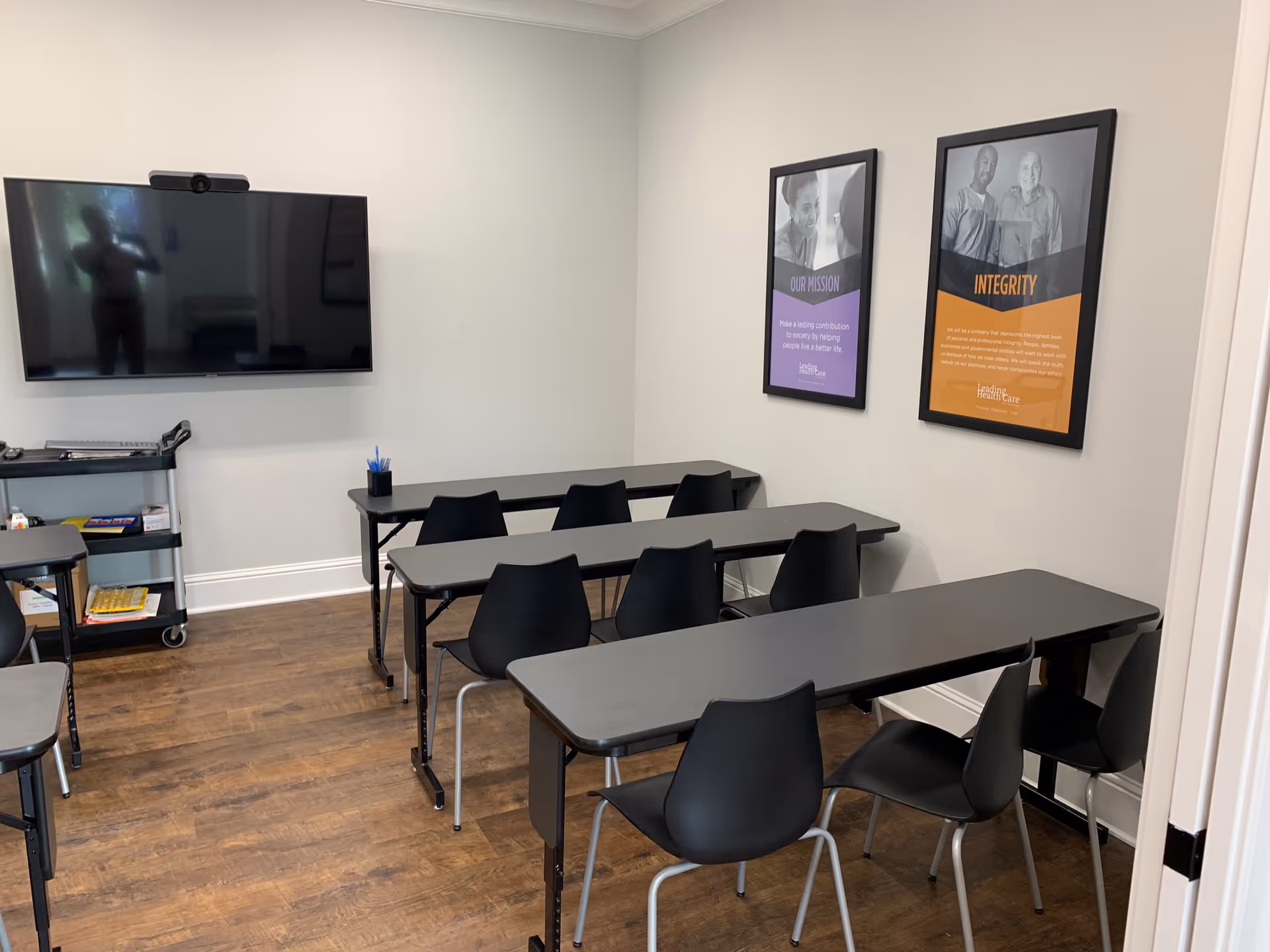 A small classroom-like training room with desks, chairs, a wall-mounted TV, and framed posters.