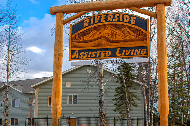 A large wooden sign reading Riverside Assisted Living and 390 Lovers Lane stands in front of a gray multi-story building with trees.