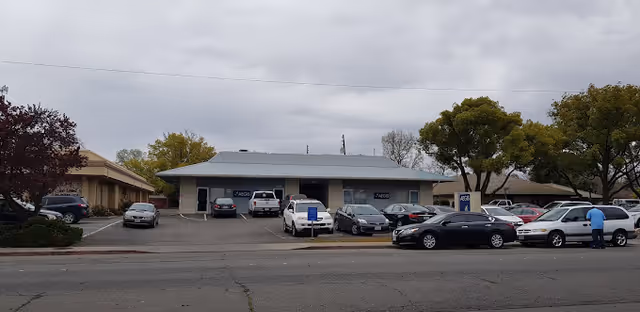 Front exterior of a single-story care facility with a parking lot of parked cars and trees under a cloudy sky.