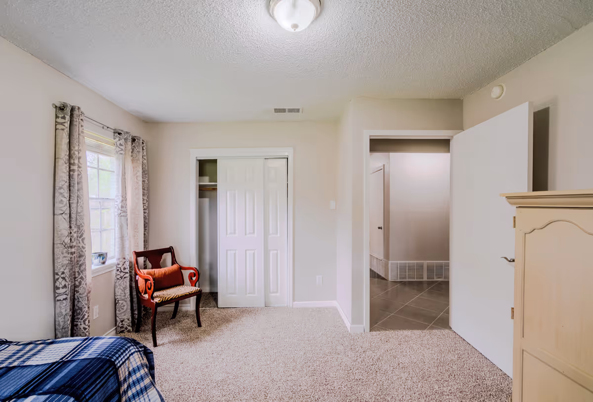 Senior living facility bedroom with window, chair, closet, and dresser.