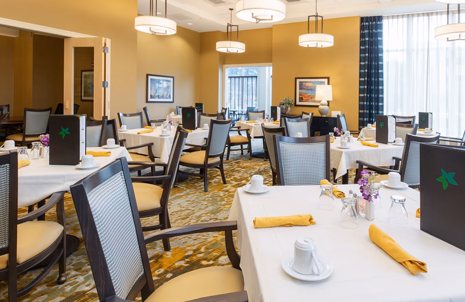 A bright, well-lit dining room with multiple tables covered in white tablecloths, each set with yellow napkins, cups, glasses, and small flower vases. The room features wooden chairs with cushioned seats and backs, warm yellow walls, framed artwork, and large windows with sheer curtains allowing natural light to fill the space.