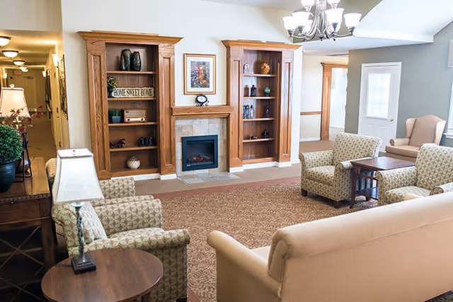 A cozy living room area with beige and patterned armchairs, a beige sofa, wooden bookshelves flanking a small electric fireplace, a chandelier overhead, and a door leading outside. The room has warm lighting and a carpeted floor.