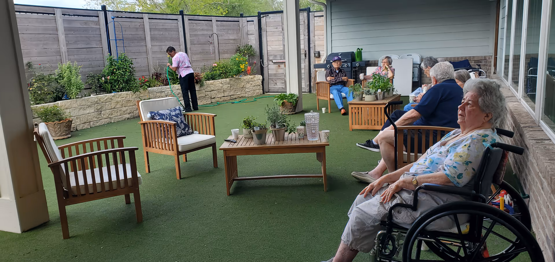 Residents relax on an outdoor patio with chairs, plants, and a wheelchair visible.