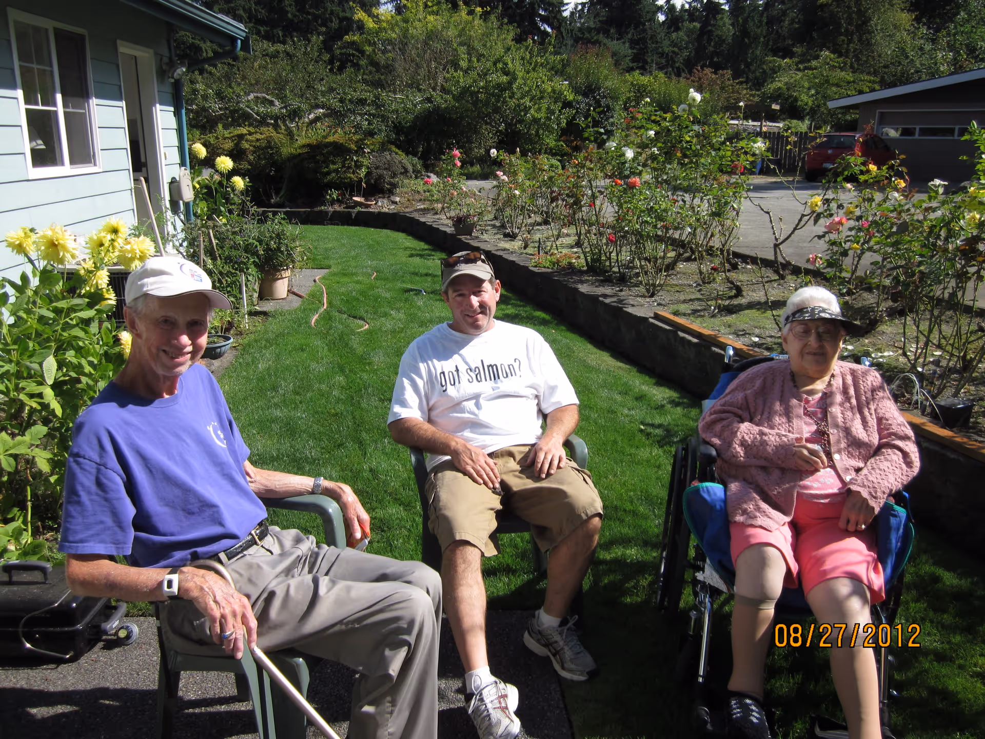 Three people sitting outside on a sunny day near a garden with flowers. One person is in a wheelchair, and the other two are seated on chairs on a grassy area next to a house and a flower bed.