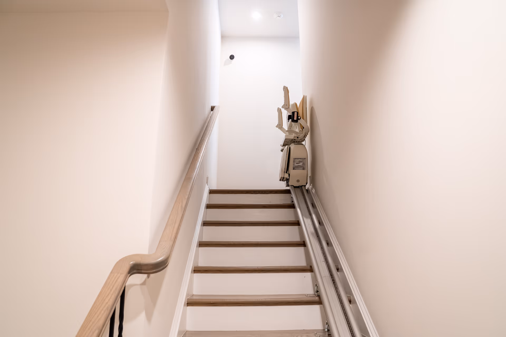 Interior stairwell with beige walls, a wooden handrail, and a stairlift along the right.
