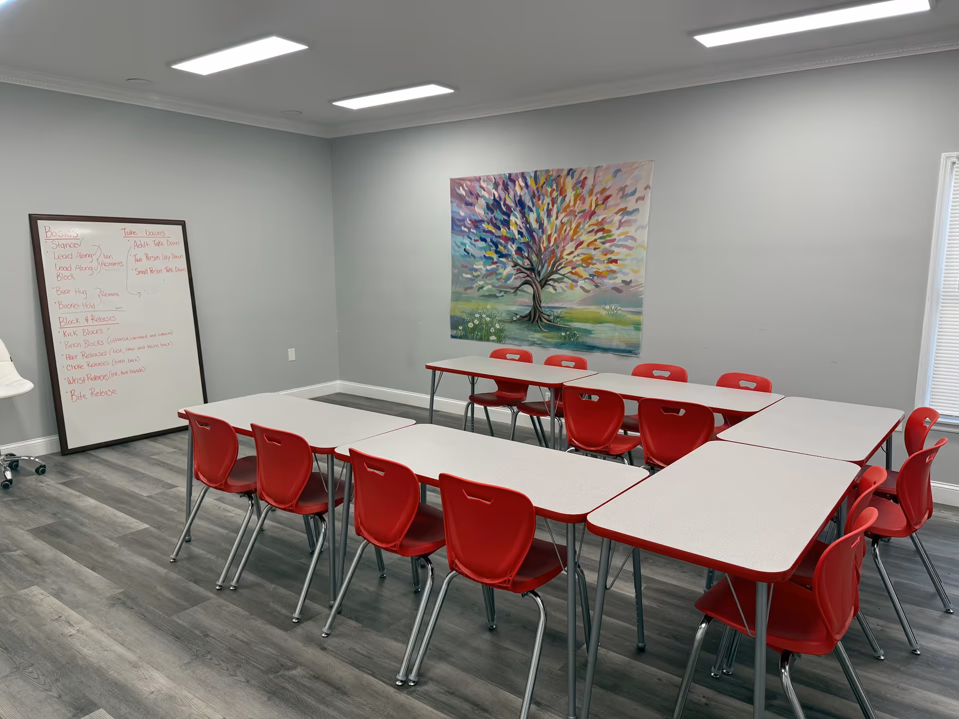 Bright classroom-style activity room with red chairs around white tables and a colorful tree painting.