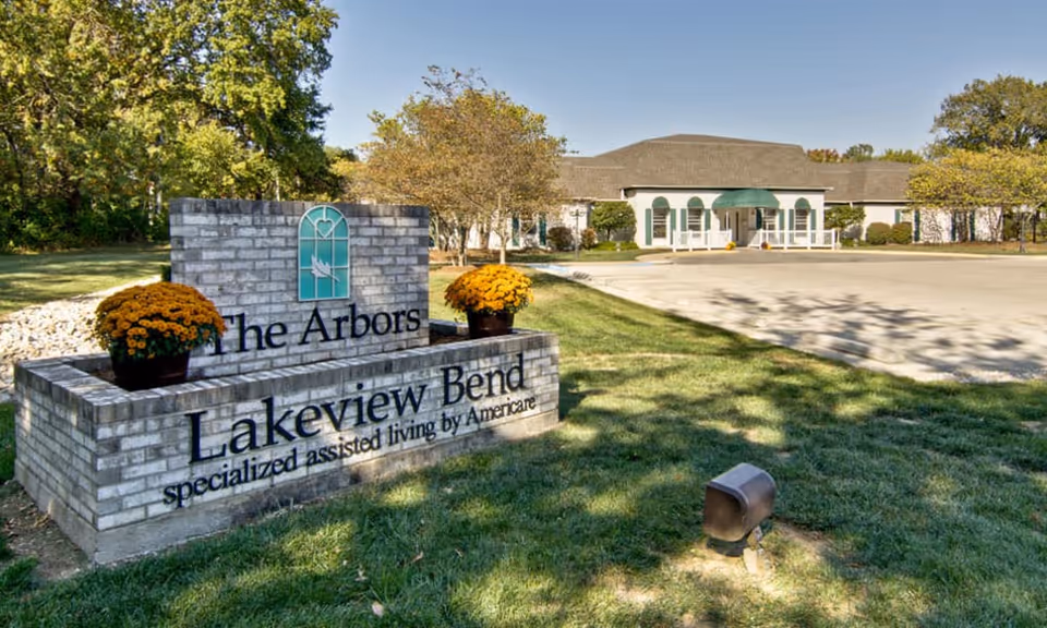 Exterior view of The Arbors at Lakeview Bend assisted living facility with a brick sign in the foreground displaying the facility's name and two pots of yellow flowers. The building is white with green shutters and an awning over the entrance, surrounded by trees and a clear blue sky.