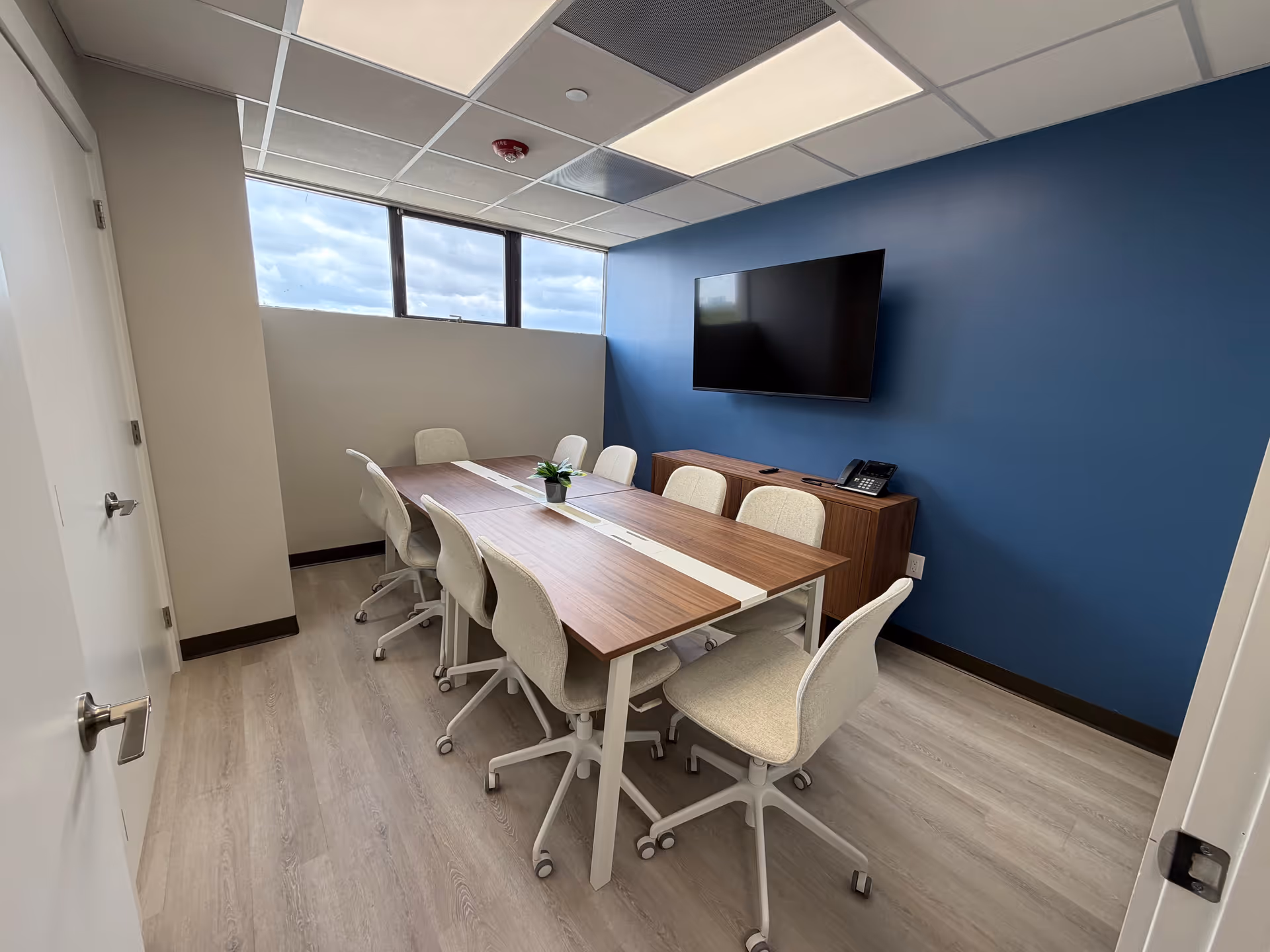 A modern conference room with a long table, light chairs, blue accent wall, and mounted TV.