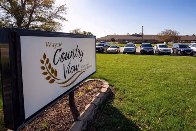 Outdoor view of Wayne Countryview Care and Rehabilitation facility sign with a landscaped area and a parking lot with several cars in front of a single-story building under a clear blue sky.
