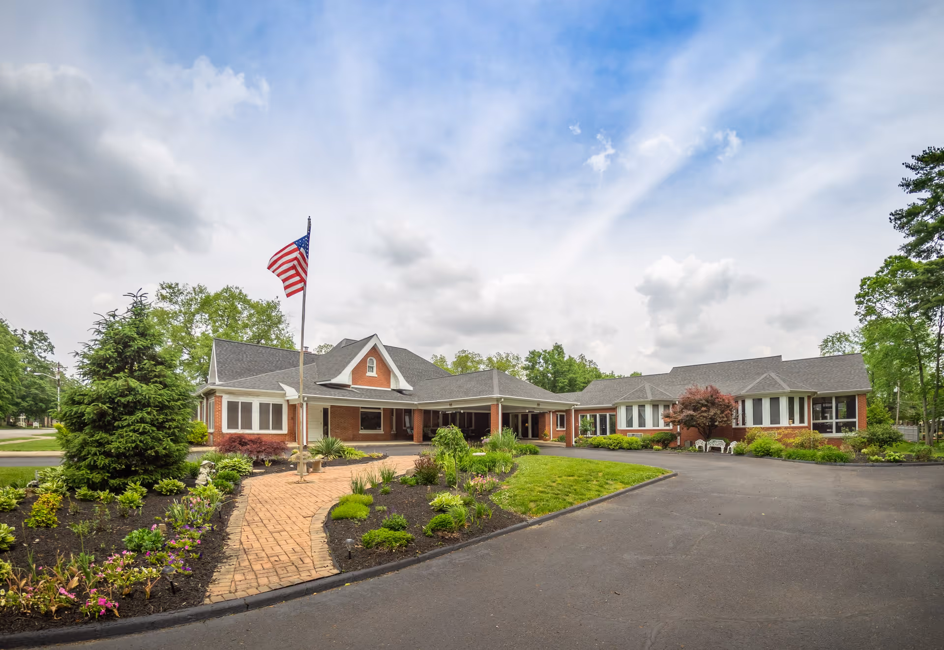 Exterior view of Charles Ford Retirement Communities of New Harmony showing a single-story brick building with a covered entrance, surrounded by landscaped gardens and a paved driveway. An American flag is flying on a flagpole near the entrance under a partly cloudy sky.