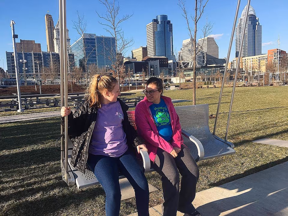 Two people sit on a park swing with a city skyline in the background.