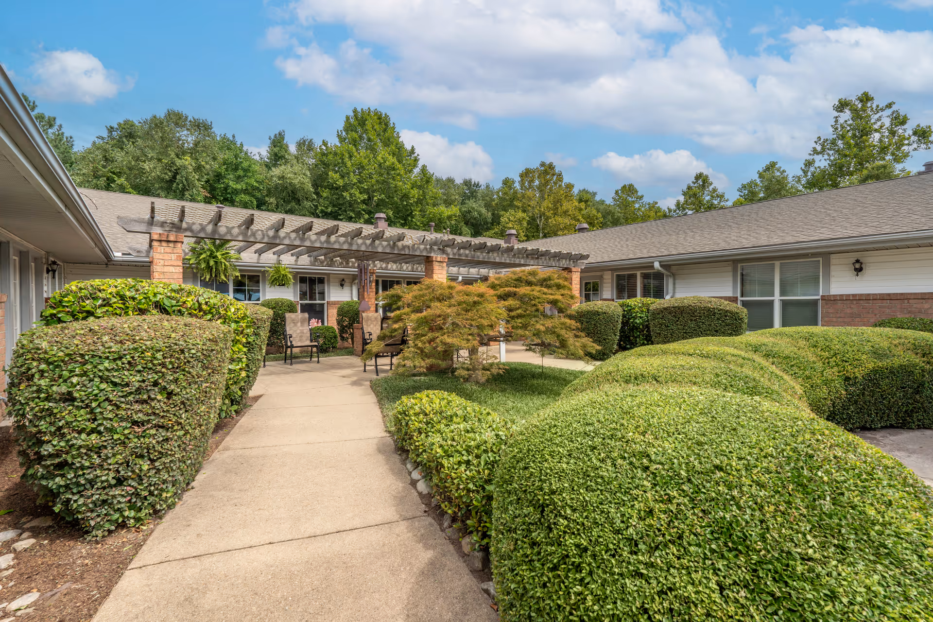 Outdoor courtyard area at Brookdale Virginia Beach featuring a concrete walkway surrounded by neatly trimmed bushes and greenery. There is a pergola with hanging plants and several chairs arranged underneath it. The building has a brick and siding exterior with windows and a shingled roof. Trees and a partly cloudy sky are visible in the background.