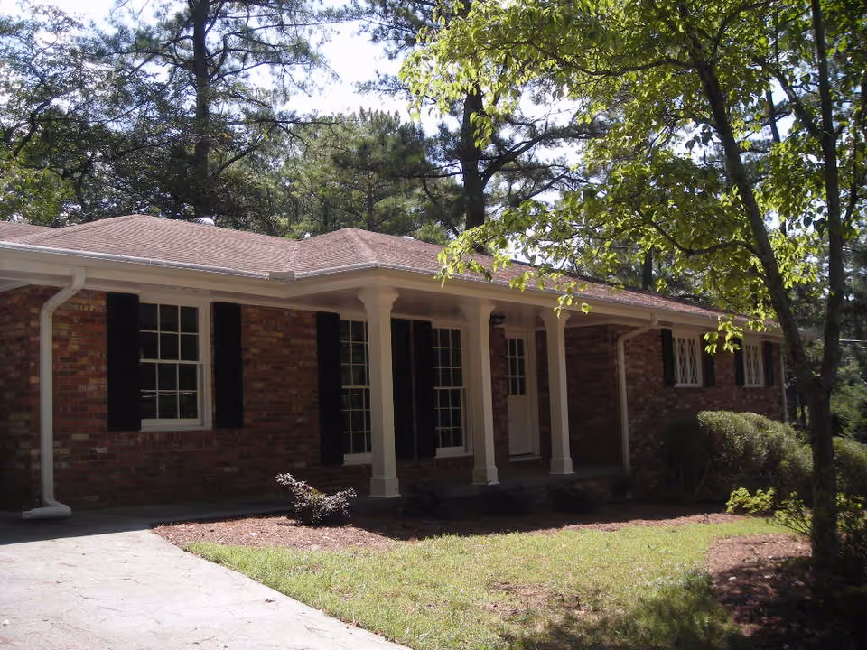 Exterior view of a single-story brick house with white columns supporting a small porch roof, black window shutters, surrounded by trees and greenery under a sunny sky.