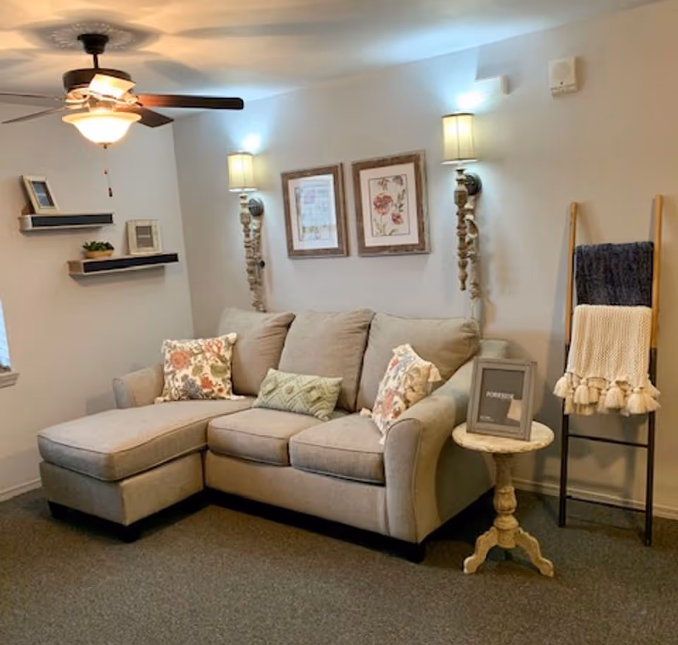 Cozy living area with a beige L-shaped sofa, floral pillows, wall sconces and framed art, a small side table and a blanket ladder.