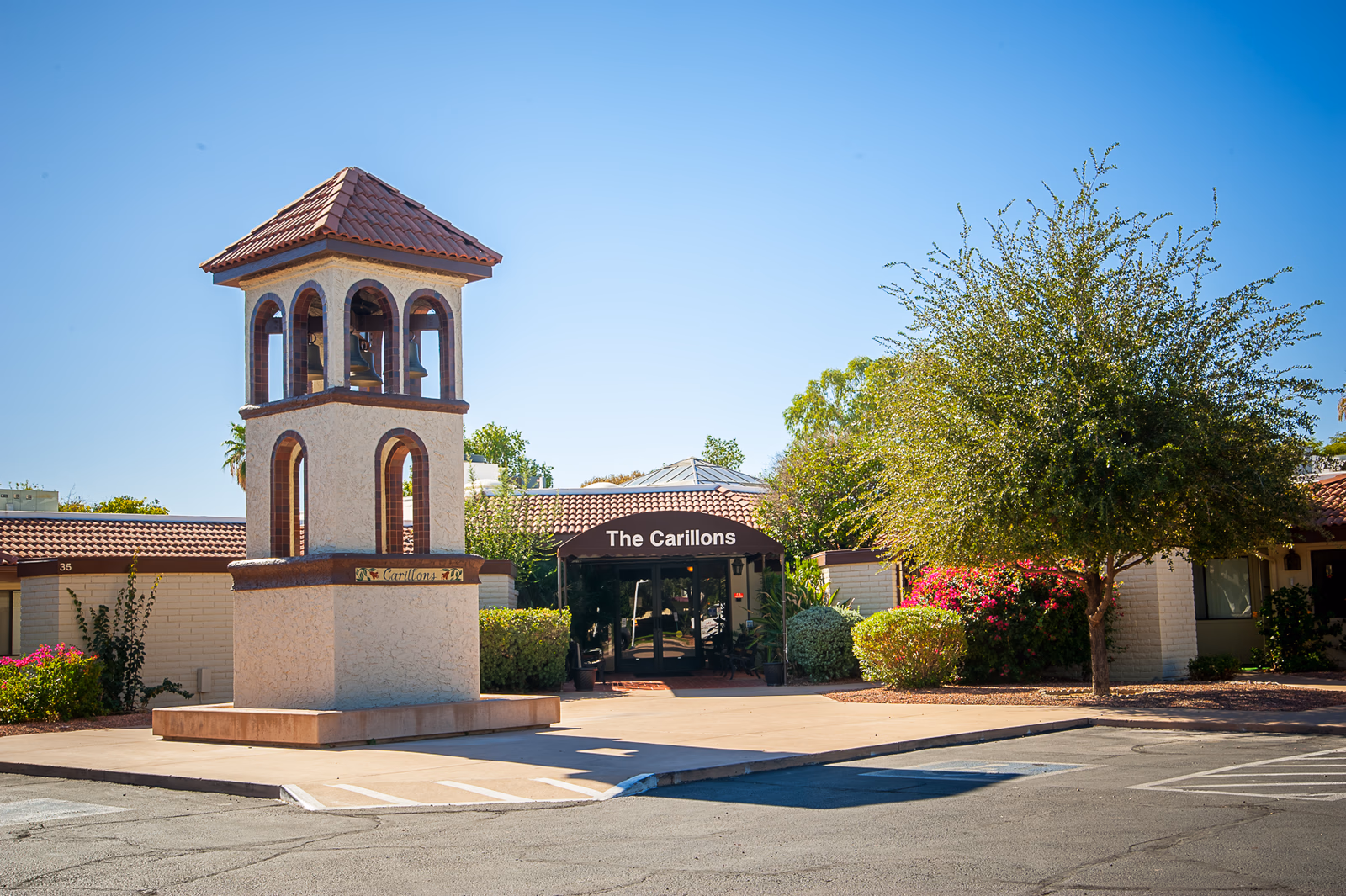 Exterior view of The Carillons Retirement Community entrance with a bell tower structure in front, surrounded by trees and bushes under a clear blue sky.