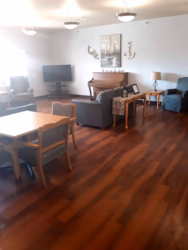 A senior living facility common area with wooden flooring, several upholstered chairs, a wooden table with chairs, a piano against the wall, a flat-screen TV on a stand, and nautical-themed wall decorations including a painting of a sailboat, a dolphin, and an anchor. There are also side tables with lamps and decorative items.
