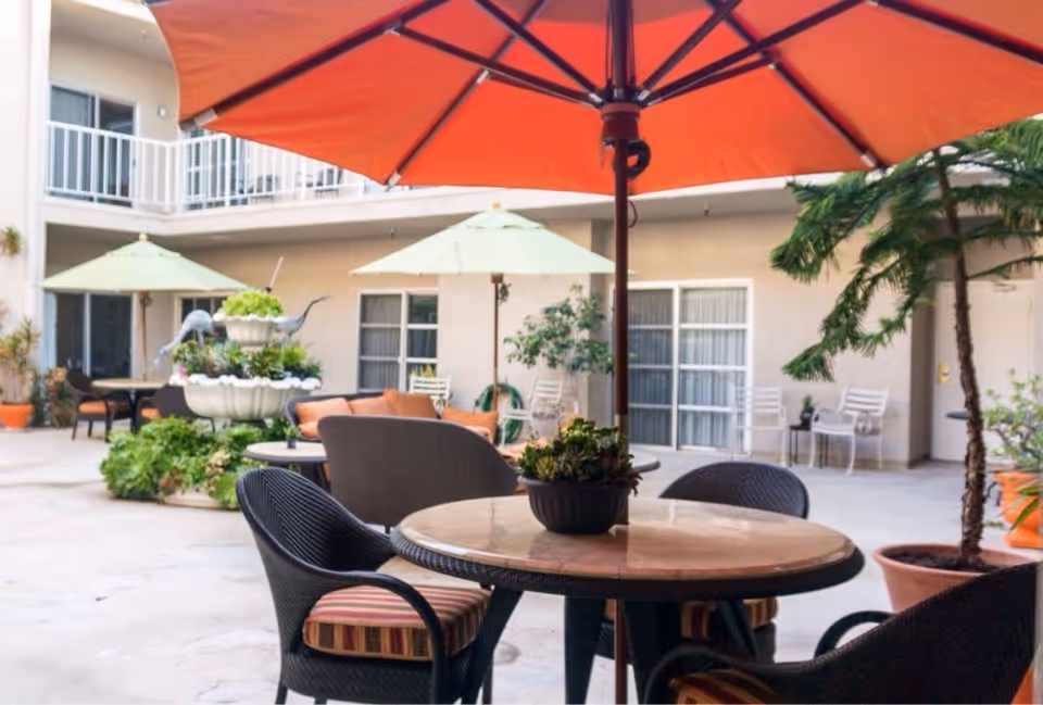Outdoor patio area with round tables and chairs under large umbrellas. There are potted plants and greenery around, with a two-story building in the background featuring balconies and windows.
