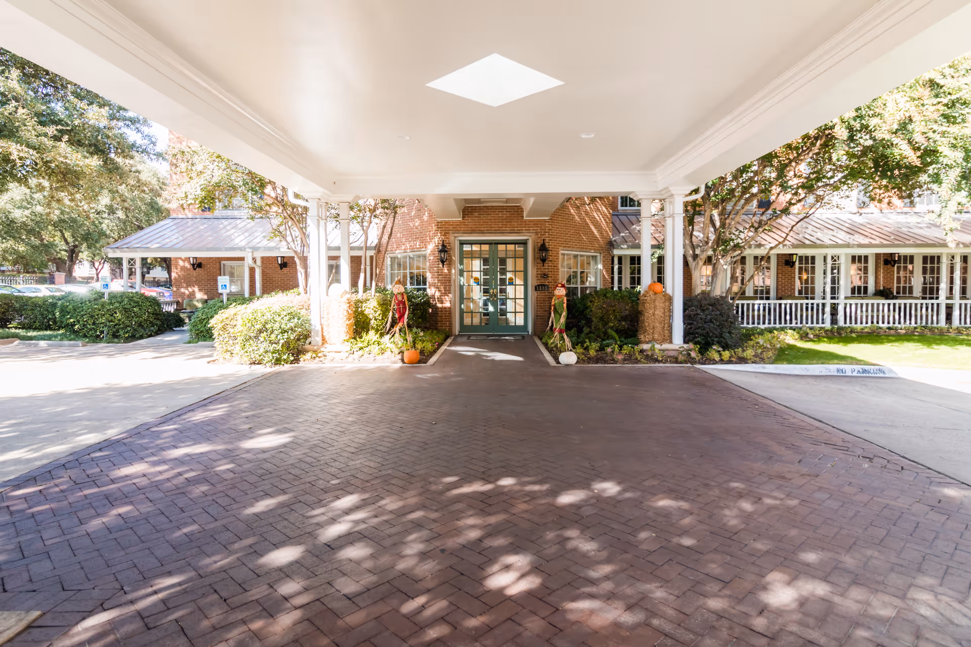 Covered entrance area of a brick building with green double doors, flanked by bushes and autumn decorations including pumpkins and scarecrows. The driveway is paved with bricks and there are trees and a parking area visible on the sides.