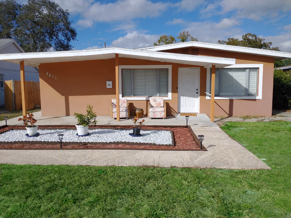 A single-story tan house with a front porch, two chairs, and planters.