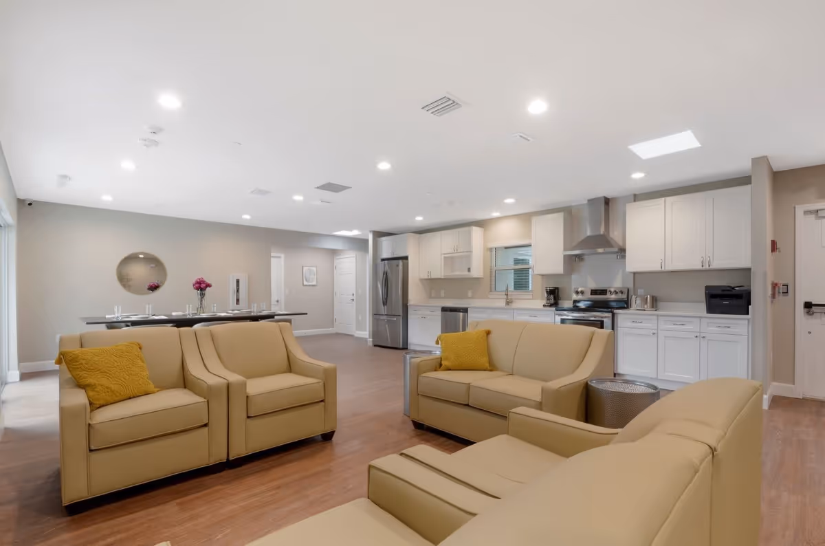 A spacious, modern common area in a senior living facility featuring beige leather sofas with yellow accent pillows arranged around a small metallic side table. In the background, there is a kitchen with white cabinets, stainless steel appliances including a refrigerator, stove, and dishwasher, and a coffee maker on the counter. To the left, a dining area with a long table set with glasses and a vase of flowers is visible. The room has wood flooring, recessed lighting, and neutral-colored walls.