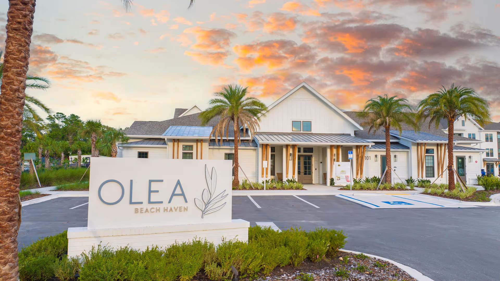 Entrance of Olea Beach Haven with a large sign, palm trees, parking spaces, and the facility building under a colorful sky.