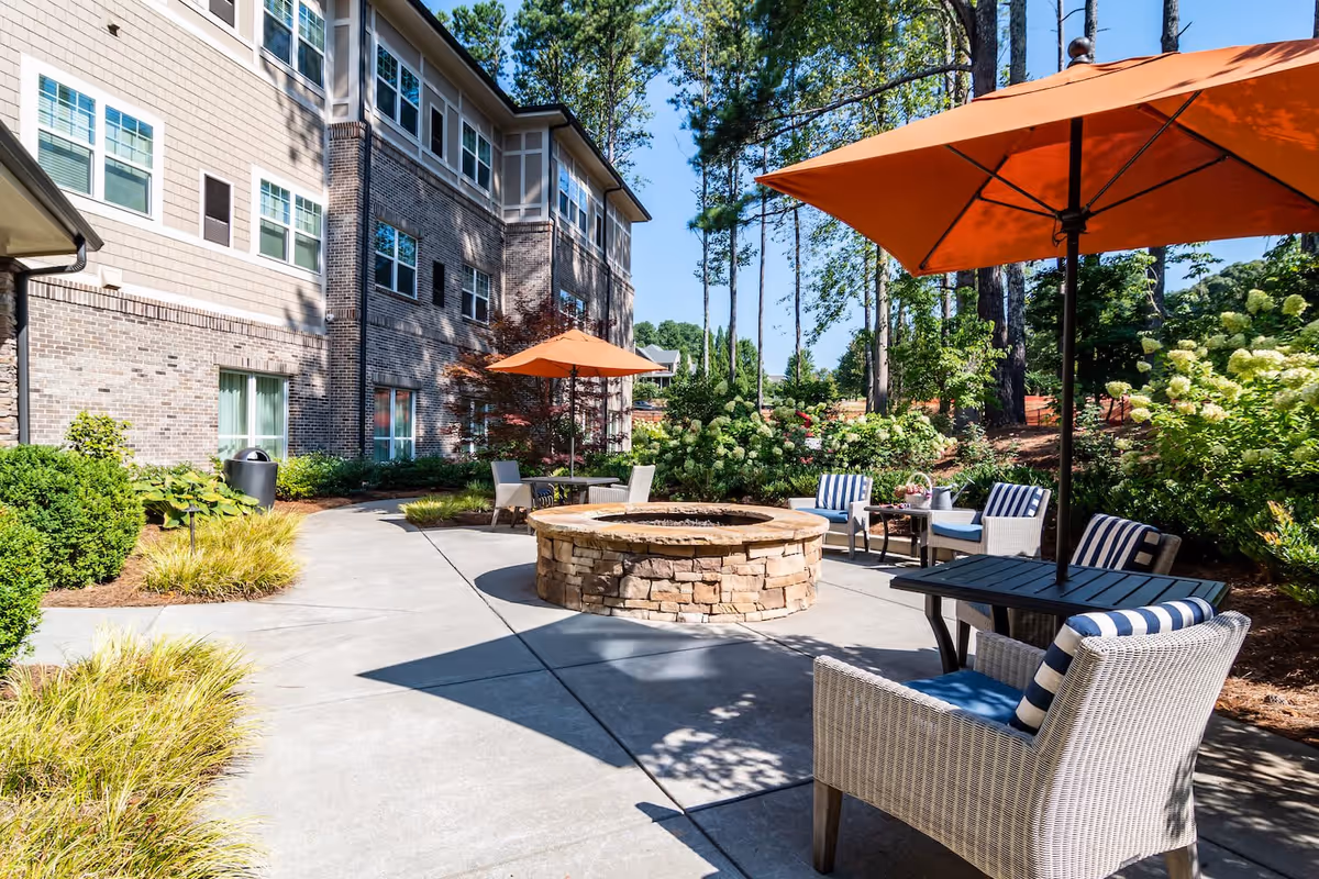 Outdoor patio area at Arbor Terrace Burnt Hickory featuring a circular stone fire pit surrounded by cushioned wicker chairs and tables with orange umbrellas. The patio is adjacent to a multi-story brick building and landscaped with green shrubs and trees under a clear blue sky.
