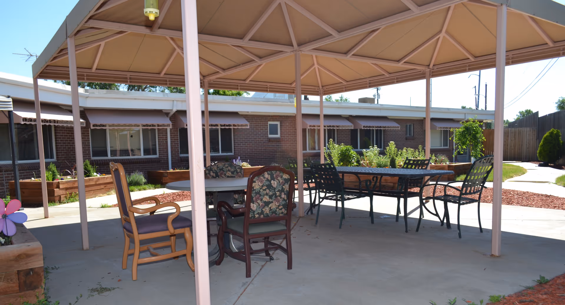 Outdoor covered seating area with a round table and several chairs, surrounded by garden beds and a brick building with windows and awnings in the background.