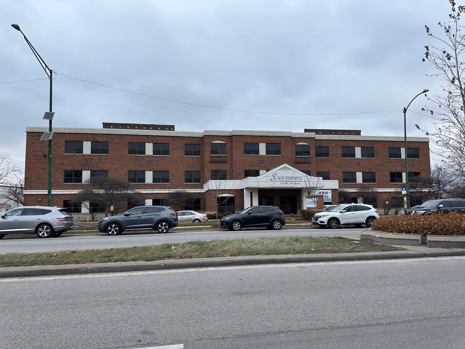 Three-story red brick nursing facility with a central covered entrance and cars parked along the street on a cloudy day.