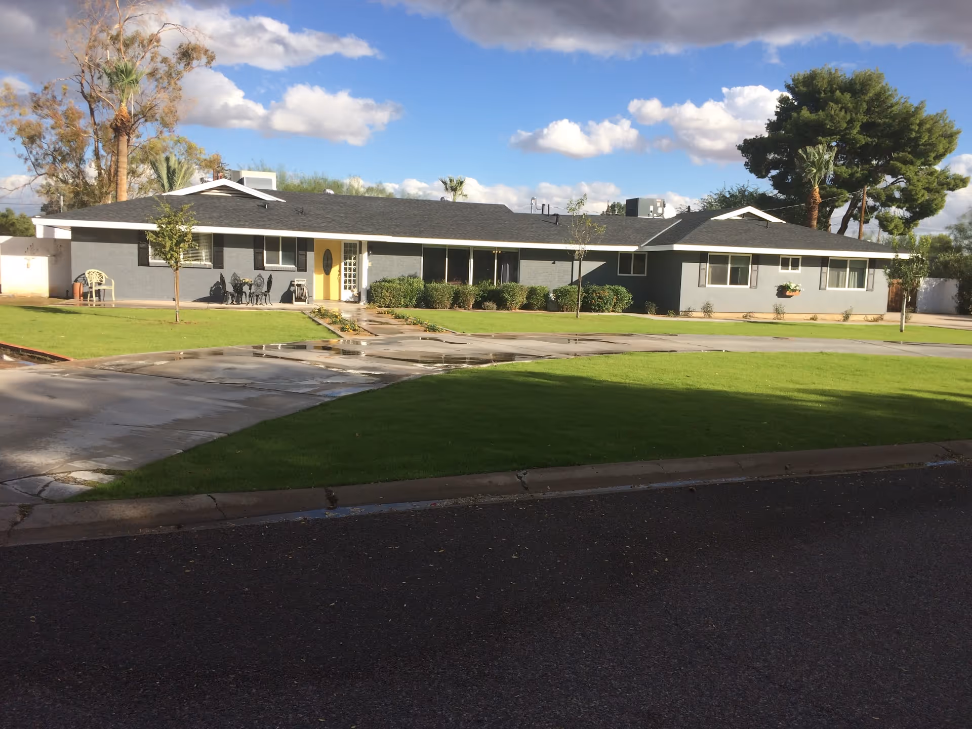 Single-story gray building with a yellow front door, surrounded by a well-maintained green lawn and a curved driveway. The sky is partly cloudy with some trees in the background.